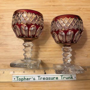 May include: Two cut-to-clear crystal goblets featuring a deep red overlay with a diamond-cut pattern on the bowl and vertical fluting at the base. Each goblet has a clear, faceted stem and a square base, positioned next to a ruler showing 10 inches.