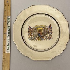 May include: A cream-colored commemorative plate with a scalloped edge, featuring a central graphic of King George V and Queen Mary for their 1935 Silver Jubilee. A wooden ruler next to the plate shows a diameter of approximately 9 inches.