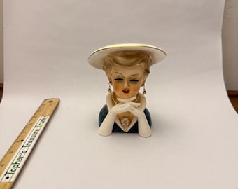 May include: A ceramic head vase depicting a figure wearing a wide-brimmed white hat with gold trim, a dark blue dress, white gloves, and a pearl necklace. The figure has blonde hair, red lips, and dangling earrings. A wooden ruler is visible nearby.