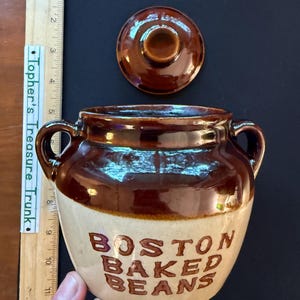 May include: A two-toned ceramic bean pot with a removable lid, featuring a dark brown glazed top and a tan base. The text "BOSTON BAKED BEANS" is embossed in brown on the front. A wooden ruler shows the pot is approximately 5.5 inches tall.