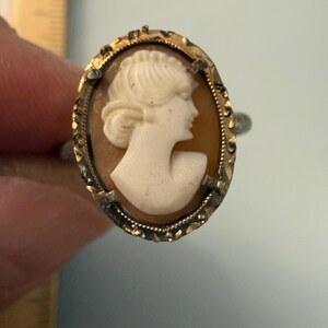 May include: An oval cameo ring featuring a white profile bust of a person against a tan background, set in a gold-toned metal frame with a textured, scalloped edge. A portion of a wooden ruler is visible on the left side of the image.