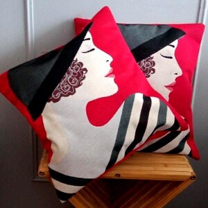 Peut inclure: Deux coussins décoratifs avec un fond rouge. Chaque coussin présente le profil d'un visage de femme en blanc, avec un chapeau noir et une lèvre rouge. Un coussin a un motif à rayures.