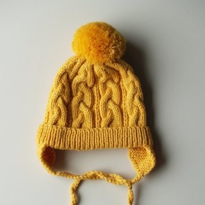 Peut inclure: Un bonnet en tricot jaune avec un pompon sur le dessus et des liens qui passent sous le menton.