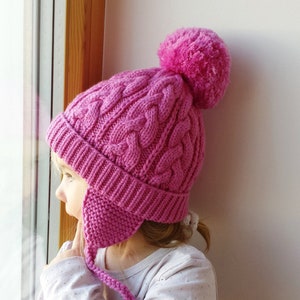 May include: A pink knitted hat with a pom pom on top and ear flaps. The hat is made with a cable knit pattern.