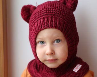 May include: A child wearing a burgundy knitted balaclava with pom-poms on top. The balaclava has a fold-down flap that covers the neck and chin. The child is looking at the camera.