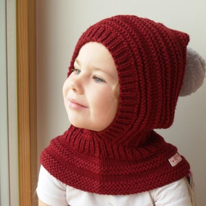Puede incluir: Una braga de cuello roja de punto con un pompón gris en la parte superior. La braga de cuello tiene una solapa abatible que cubre el cuello y la barbilla. La braga de cuello está hecha de un material suave y cálido, perfecto para mantenerse caliente en climas fríos.