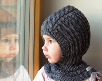 May include: A gray knitted balaclava with a cable knit pattern. The balaclava is pulled up over the child's head, covering their ears and neck. The child is looking out a window.