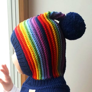 Puede incluir: Un gorro de punto azul con un patrón de rayas de arcoíris y un pompón azul en la parte superior. El gorro tiene un cuello de punto azul a juego.