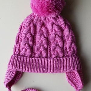 May include: A pink knitted hat with a pom-pom on top and ear flaps. The hat has a cable knit pattern and a long tie.