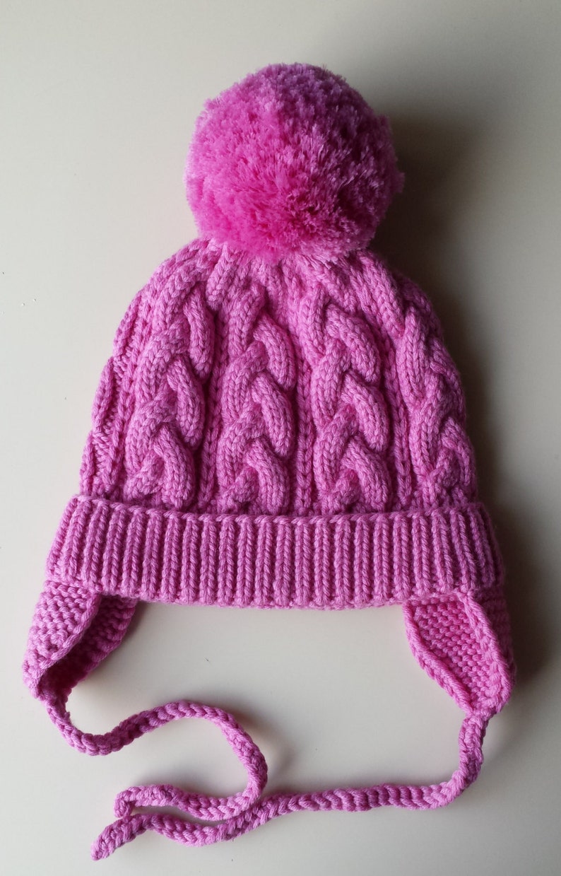 May include: A pink knitted hat with a pom-pom on top and ear flaps. The hat has a cable knit pattern and a long tie.