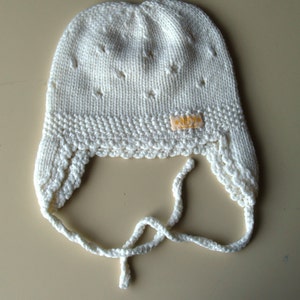 May include: White knitted baby bonnet with a tie. The bonnet has a small label that says "*NSYNC* Handmade"