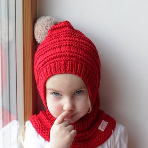 Puede incluir: Un gorro de punto rojo con un pompón en la parte superior. El gorro tiene una solapa que se pliega hacia abajo para cubrir el cuello y la barbilla. El gorro lo lleva un niño con el pelo rubio.
