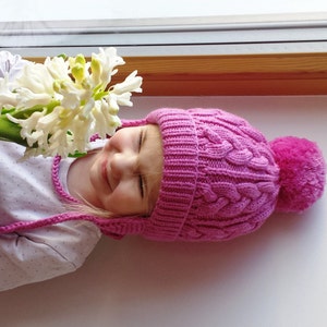 May include: A young girl wearing a pink knitted hat with a pom-pom on top, looking at a white hyacinth flower in a pot.