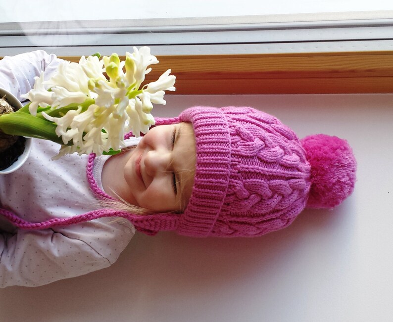 May include: A young girl wearing a pink knitted hat with a pom-pom on top, looking at a white hyacinth flower in a pot.