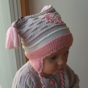 May include: A knitted baby hat with a pink and white striped design and a pink flower applique. The hat has a long pink tassel and ear flaps.