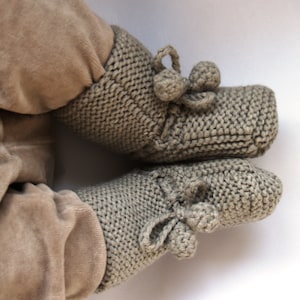 Può includere: Un paio di scarpine per neonati in maglia di colore marrone chiaro con un fiocco e dei pon pon.