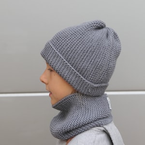 Puede incluir: Un conjunto de gorro y braga de cuello de punto gris. El gorro tiene un borde doblado y una parte superior redondeada. La braga de cuello es un tubo que se ajusta alrededor del cuello. Ambos artículos están hechos de un tejido de punto grueso y texturizado. El conjunto está diseñado para la calidez y la comodidad.