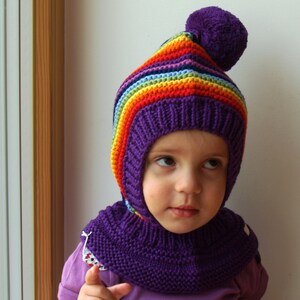 Puede incluir: Un niño lleva un gorro de punto morado con un patrón de rayas de arcoíris y un cuello de punto morado a juego. El gorro tiene un pompón morado esponjoso en la parte superior.