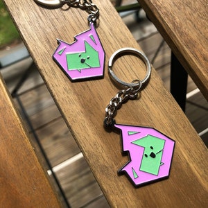 May include: Two pink and green enamel keychains with silver key rings and chains. Each keychain features a cartoon dog with a green face and black eyes.