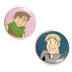 Könnte beinhalten: Zwei runde Ansteckbuttons mit Cartoon-Illustrationen. Ein Button hat einen rosa Hintergrund mit einem Cartoon-Mann mit grünem Schal. Der andere Button hat einen blauen Hintergrund mit einem Cartoon-Mann mit blonden Haaren.