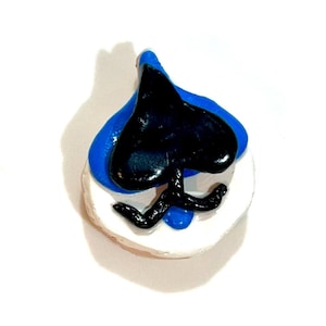 Könnte beinhalten: Ein handgefertigter Pik-Ass-Anhänger. Der Anhänger hat eine schwarze Pikform über einem blauen Tropfendesign, das auf einer weißen, kreisförmigen Basis angeordnet ist. Der Anhänger ist wahrscheinlich aus Ton oder einem ähnlichen Material gefertigt.