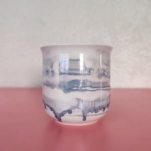 Puede incluir: Una taza de cerámica blanca con un patrón horizontal abstracto de trazos de pintura azul grisáceo que parecen gotear hacia abajo. La taza tiene un acabado liso y brillante, con un borde ligeramente ensanchado y una forma cilíndrica sin asa.