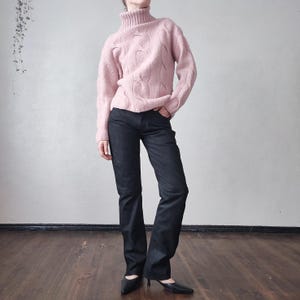 May include: A pale pink cable-knit turtleneck jumper worn with dark denim jeans. The jeans have a subtle flare. Black pointed-toe heels complete the outfit, creating a casual yet stylish look.