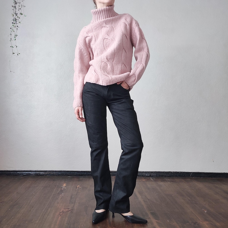 May include: A pale pink cable-knit turtleneck jumper worn with dark denim jeans. The jeans have a subtle flare. Black pointed-toe heels complete the outfit, creating a casual yet stylish look.
