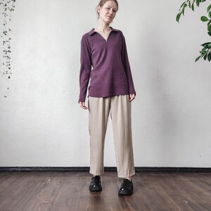 May include: A woman wearing a long-sleeved, ribbed, plum-colored shirt with a collared neckline and a zipper. She is also wearing beige, wide-legged pants and black shoes. The outfit is set against a white wall and a dark wooden floor.