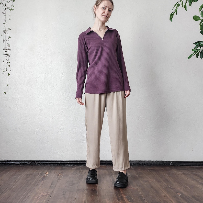 May include: A woman wearing a long-sleeved, ribbed, plum-colored shirt with a collared neckline and a zipper. She is also wearing beige, wide-legged pants and black shoes. The outfit is set against a white wall and a dark wooden floor.
