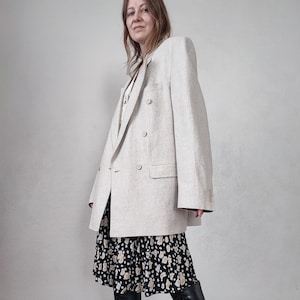 May include: A light beige double-breasted blazer with a classic design. The blazer has two rows of buttons and a tailored fit. The model is wearing a black and white patterned dress and black boots. The blazer is made of a textured fabric.