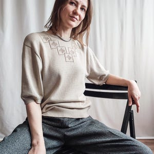 Puede incluir: Una mujer lleva un suéter beige con un patrón geométrico y pantalones grises sentada en una silla negra.