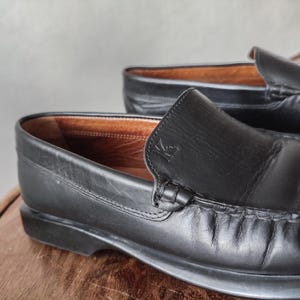 K&ouml;nnte beinhalten: Nahaufnahme eines Paares schwarzer Leder-Loafers. Die Schuhe haben ein klassisches Design mit runder Spitze und einem Schlupfstil. Die Innenseite ist hellbraun. Die Schuhe stehen auf einer Holzoberfl&auml;che.