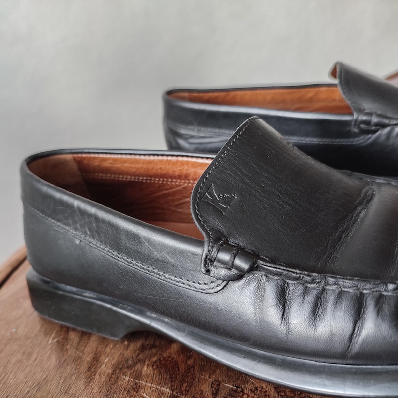 K&ouml;nnte beinhalten: Nahaufnahme eines Paares schwarzer Leder-Loafers. Die Schuhe haben ein klassisches Design mit runder Spitze und einem Schlupfstil. Die Innenseite ist hellbraun. Die Schuhe stehen auf einer Holzoberfl&auml;che.