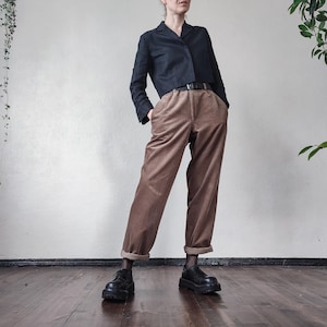 Könnte beinhalten: Ein dunkelblauer, gecroppter Blazer kombiniert mit einer hoch taillierten Cordhose mit weitem Bein in einem warmen Braunton. Das Outfit wird durch einen schwarzen Gürtel und klobige schwarze Schuhe ergänzt. Die Hose ist an den Knöcheln umgeschlagen.