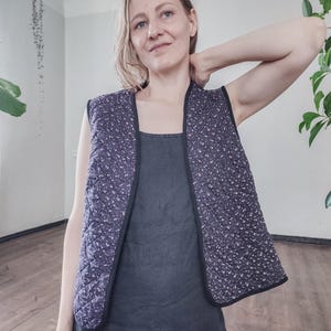 May include: A quilted vest in a dark blue fabric with a white floral pattern. The vest has a black trim and a sleeveless design. The model is wearing a dark blue dress underneath the vest. The vest is a stylish layering piece.