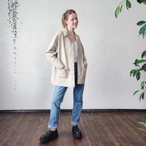May include: A beige jacket with a collared neckline, button closure, and front pockets. The jacket is paired with blue jeans and black shoes. The outfit is casual and suitable for everyday wear.