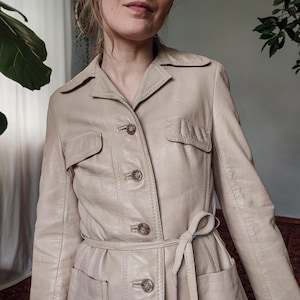 Puede incluir: Chaqueta de cuero beige claro con cuello, cierre de botones y dos bolsillos delanteros. La chaqueta tiene un cinturón a juego y es de estilo vintage. La chaqueta es de color neutro.