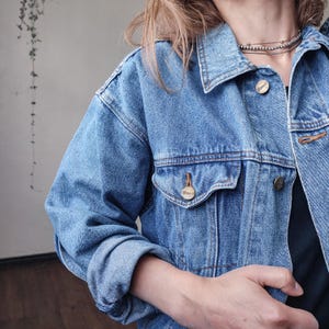 May include: A light blue denim jacket with a classic collar, button closures, and front pockets. The jacket has a slightly oversized fit and features gold-colored buttons and rivets. The sleeves are cuffed, and the jacket is worn over a dark top.