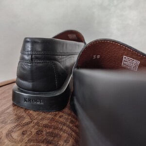 K&ouml;nnte beinhalten: Nahaufnahme von zwei schwarzen Leder-Loafern. Die Sohle eines Schuhs ist mit dem Markennamen "KROLL" versehen. Die Innenseite des anderen Schuhs zeigt die Gr&ouml;&szlig;e "38". Die Schuhe stehen auf einer Holzoberfl&auml;che.