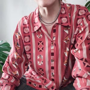 Może przedstawiać: Czerwona koszula zapinana na guziki z geometrycznym wzorem. Koszula ma pionowe paski i powtarzające się kształty w odcieniach czerwieni, bieli i brązu. Długie rękawy mają mankiety zapinane na guziki, a kołnierz jest widoczny.