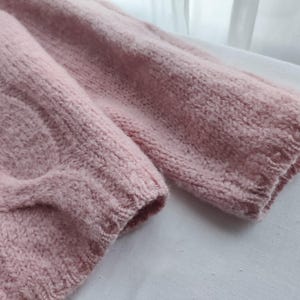 May include: Close-up of a pale pink knitted sweater. The sleeves are visible, showing the textured knit and ribbed cuffs. The soft colour and cosy texture suggest warmth and comfort, ideal for cooler weather.