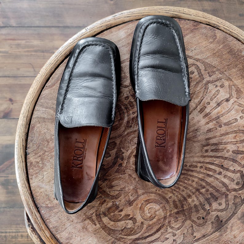 K&ouml;nnte beinhalten: Ein Paar schwarze Leder-Loafer mit quadratischer Zehenpartie. Die Schuhe haben einen Slip-on-Stil und tragen den Markennamen "KROLL" auf der Innensohle. Die Loafer liegen auf einer Holzoberfl&auml;che mit einem dekorativen Muster.