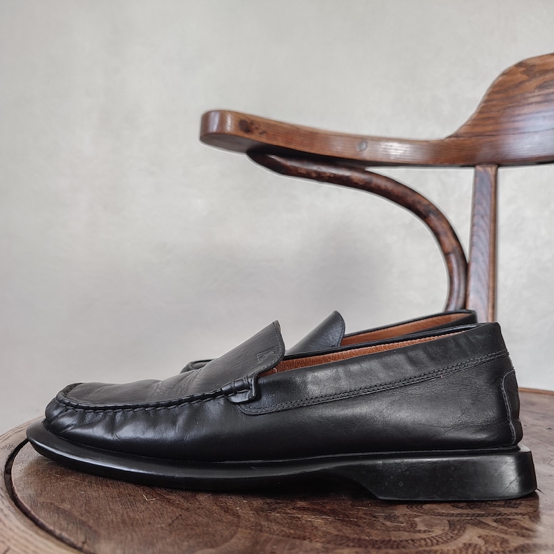 K&ouml;nnte beinhalten: Ein Paar schwarze Leder-Loafer mit runder Spitze und Slip-on-Design. Die Schuhe werden auf einem Holzstuhl pr&auml;sentiert, was ihren klassischen Stil und ihre hochwertige Verarbeitung unterstreicht. Die Loafer haben ein gen&auml;htes Detail um den Vorderfu&szlig;.