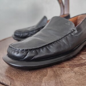 K&ouml;nnte beinhalten: Ein Paar schwarze Leder-Loafer. Die Schuhe haben eine runde Spitze, ein Slip-on-Design und eine Nahtdetail am Vorderblatt. Die Sohlen sind schwarz und leicht erh&ouml;ht. Die Schuhe stehen auf einer Holzoberfl&auml;che.