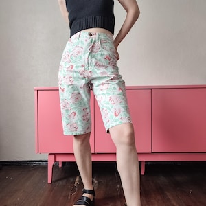 May include: High-waisted denim shorts featuring a light mint green base color with a repeating pattern of soft pink roses and green leaves. The shorts have a classic five-pocket design, a button fly closure, and a knee-length cut with a straight leg.