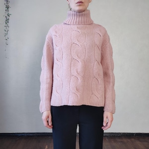 May include: A pale pink cable-knit turtleneck jumper. The jumper features a high, ribbed collar and a textured design. The person is wearing black trousers. The jumper is a warm, comfortable garment.