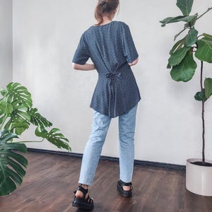Puede incluir: Una blusa azul marino con lunares blancos, combinada con vaqueros azul claro y sandalias negras. La blusa tiene mangas cortas y un lazo en la espalda. El conjunto se lleva en una habitación con plantas y suelo de madera.