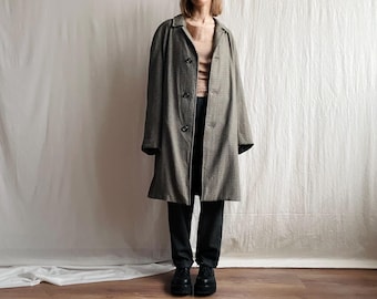 May include: A long, button-down coat in a black and tan checkered pattern. The coat has a classic collar and is worn over a neutral-colored top and black pants. The outfit is completed with black shoes.