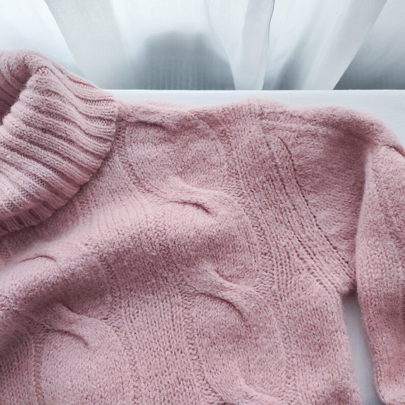 May include: A close-up of a pale pink cable-knit jumper. The jumper features a roll neck and a textured cable design across the front and sleeves. The soft knitwear is a cosy and comfortable garment.